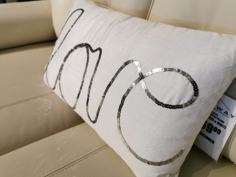 The Ashley Newry 'Love' Pillow features a sequin message and sits on a beige leather sofa.