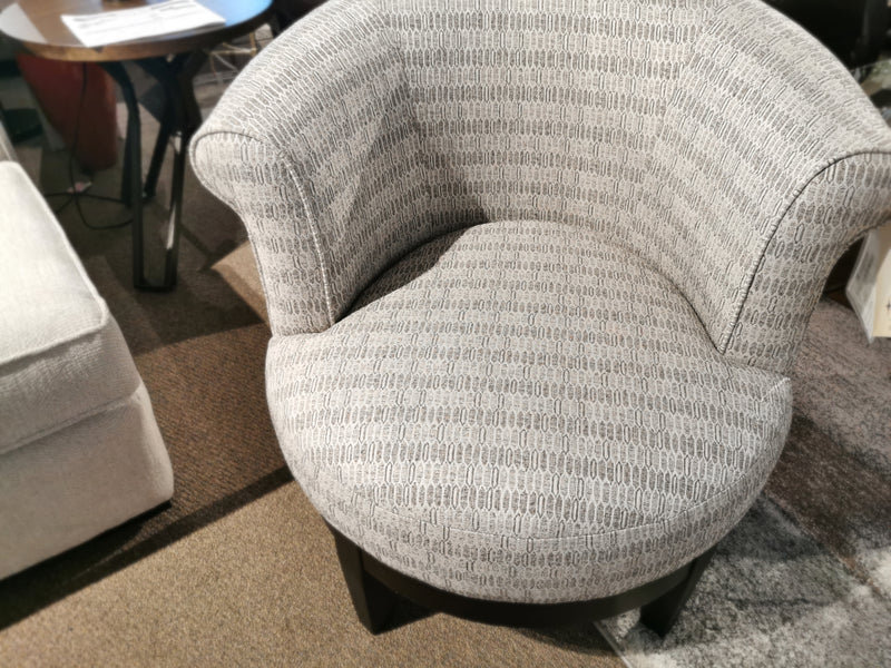 The Attica Swivel Chair by Best, featuring a gray upholstery with a textured pattern, is placed on a brown carpet next to a small dark side table, adding a touch of cottage style charm. This accent chair perfectly complements cozy living spaces.