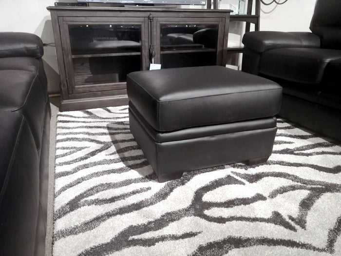 A Colorado Leather Ottoman by Leather Craft on a zebra-patterned rug between two black sofas, with a dark wood cabinet behind.