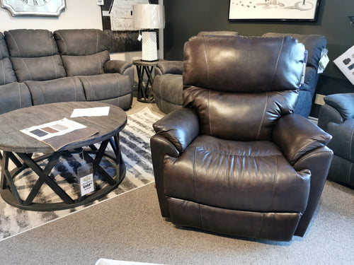In the furniture showroom, the 724 Trouper Leather Rocker Recliner by La-Z-Boy stands proudly with its sleek design in rich brown leather, flanked by gray couches and complemented by a round wooden coffee table. It promises style while offering essential lumbar support for ultimate comfort.