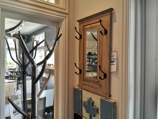 In the store, Woodcraft's 712 Doorganizer is elegantly displayed. This wooden wall-mounted mirror with coat hooks features a vintage grey stain and is priced at $199.99. Alongside it, a partially visible coat rack offers smart 2-D space storage solutions while complementing the functional and stylish design of this piece.