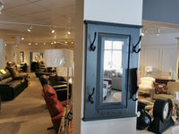 A furniture store showroom features a selection of sofas and chairs, with a wall adorned with mirrors and hooks. Prominently displayed in the foreground is the 712 Doorganizer by Woodcraft, expertly crafted with a vintage grey stain for stylish 2-D space storage.